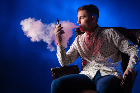 A man smokes a vape. Guy is looking at the vape device. A young man blows a cloud of steam. Smoking...