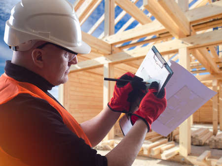 A builder next to wooden house. Timber frame behind architect. Man draw construction drawings. Architect with blueprints at construction site. Builder in working uniform. career in architectureの写真素材