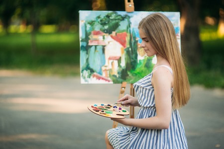The blonde girl paints a painting on the canvas with the help of paints. A wooden easel keeps the picture. summerの写真素材