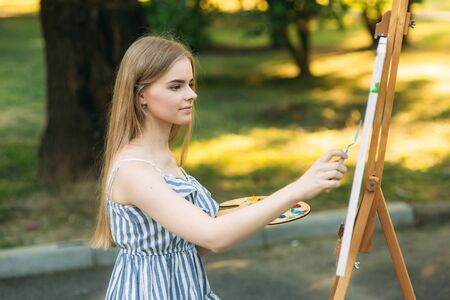 The blonde girl paints a painting on the canvas with the help of paints. A wooden easel keeps the picture. summerの写真素材