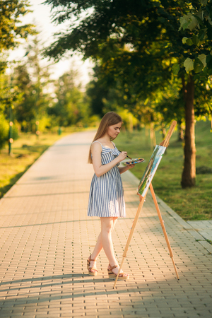 Beautiful girl draws a picture in the park using a palette with paints and a spatula. Easel and canvas with a picture. Summer is a sunny day, sunset. dayの写真素材