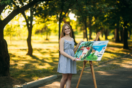 The blonde girl paints a painting on the canvas with the help of paints. A wooden easel keeps the picture. summerの写真素材