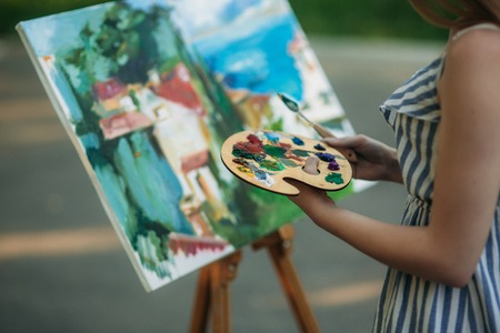The blonde girl paints a painting on the canvas with the help of paints. A wooden easel keeps the picture. summerの写真素材