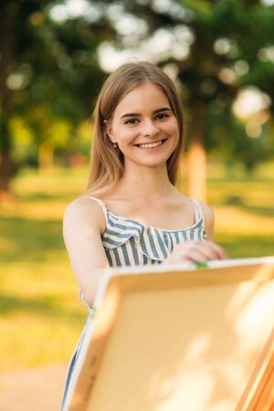 Beautiful girl draws a picture in the park using a palette with paints and a spatula. Easel and canvas with a picture.の写真素材