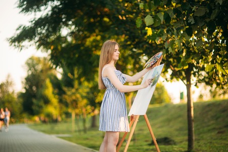 Beautiful girl draws a picture in the park using a palette with paints and a spatula. Easel and canvas with a picture.の写真素材