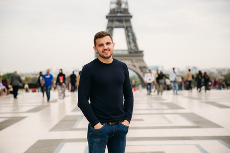 A young man wearing a dark blue jacket is standing on the background of the Eiffel Tower. Sunny weather is autumnの写真素材