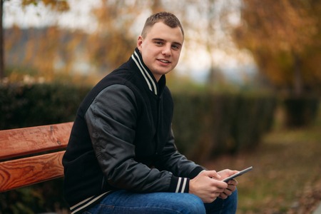 A student in a black jacket sits in a park on the bench and use tablet. Handsome boyの写真素材