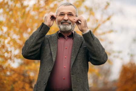 A handsome elderly man wearing glasses is using a phone. Walk in the park in autumn