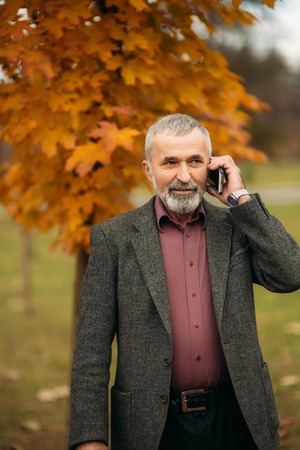 A handsome elderly man wearing glasses is using a phone. Walk in the park in autumnの写真素材