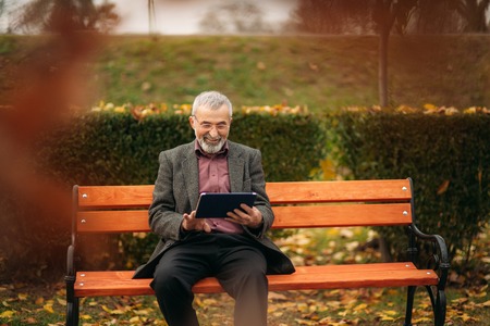 Grandpa use a tablet sitting in the pakr on the benchの写真素材