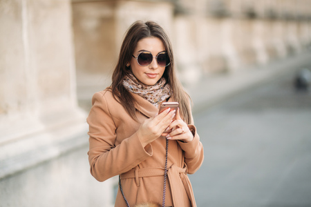 Stylish girl in a brown coat and a scarf with glasses. Using a phone in paris france. Makeupの写真素材