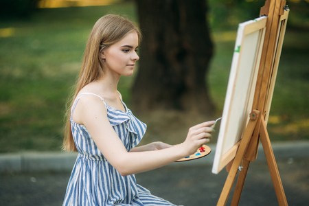 Beautiful girl draws a picture in the park using a palette with paints and a spatula.の写真素材