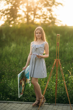 Beautiful girl draws a picture in the park using a palette with paints and a spatula. Easel and canvas with a picture.の写真素材