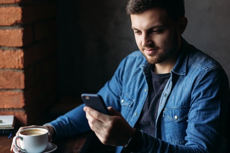 Bearded man in a denim shirt using phone inside a restaurant while drinking a coffeeの写真素材