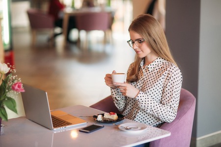 Girl eating cake in a cafe. Blonde in polka-dot shirt with laptopの写真素材