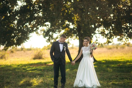 Stylish couple of happy newlyweds walking in field on their wedding day with bouquet. In the middle of the field ther is a big treeの写真素材