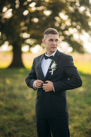 Groom makes a button on his jacket. standing in the middle of field in the background of big treeの写真素材