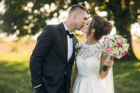 Stylish couple of happy newlyweds walking in field on their wedding day with bouquet. In the middle of the field ther is a big treeの写真素材