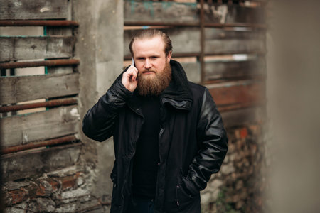 A bearded man in a black leather jacket stands outside and poses for a photographer and using a...