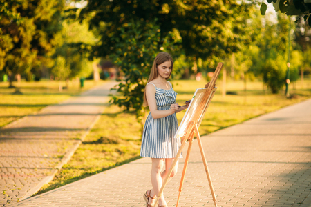 Beautiful girl standing in the park and draws a picture using a palette with paints and a spatula.の写真素材