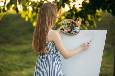 Beautiful girl standing in the park and draws a picture using a palette with paints and a spatulaの写真素材