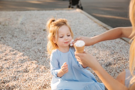 Beautiful little girl in a blue dress eating an ice cream, Mum helps and wipes her mouthの写真素材