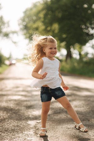 A cheerful adorable little girl with blond hair dances and jumps through the alley on a warm summer...