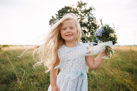 Little girl in sky blue dress with wreath and bouquet stand in field in front of big tree