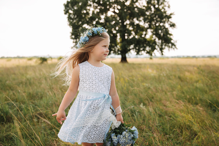 Little girl in sky blue dress with wreath and bouquet stand in field in front of big treeの写真素材