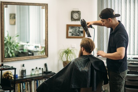 Master cuts hair and beard of men in the barbershop and uses a hair dryerの写真素材