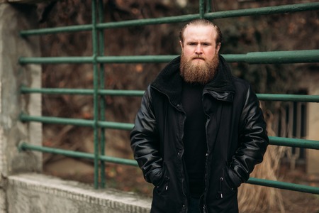 A bearded man in a black leather jacket stands outside and poses for a photographerの写真素材
