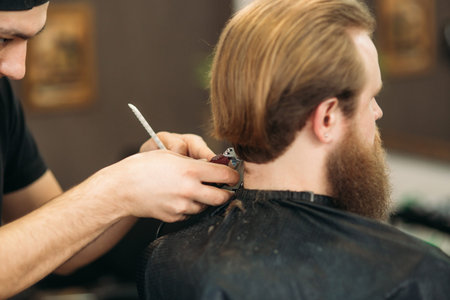 The hands of young barber making haircut to attractive bearded man in barbershop
