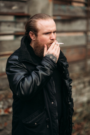 A bearded man in a black leather jacket stands outside and poses for a photographer. Smokes sigaretの写真素材