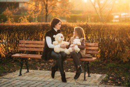 Mother and little daughter is sitting on a bench in the park. Holds a teddy bearの写真素材