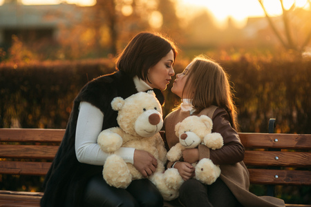 Mother and little daughter is sitting on a bench in the park. Holds a teddy bearの写真素材
