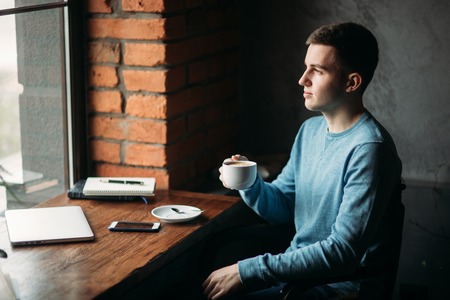 Handsome boy drinking coffee in cafe. He sits near the window with laptop and notebookの写真素材