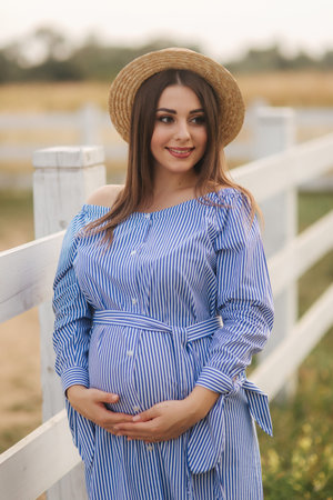 Beautiful pregnant woman in blue dress and knitted hat stand near the fance in the farm. She put...