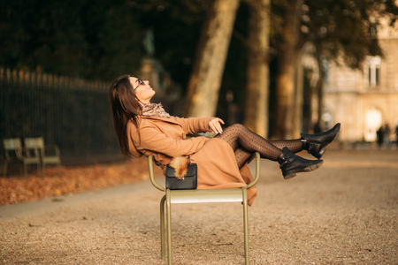A young girl in a brown coat sits in the middle of the park on a khaki stool.の写真素材