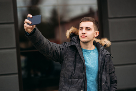 Man in a gray jacket is standing outside using a phone and making selfieの写真素材