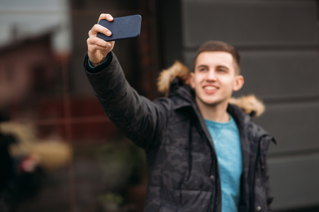 Man in a gray jacket is standing outside using a phone and making selfieの写真素材