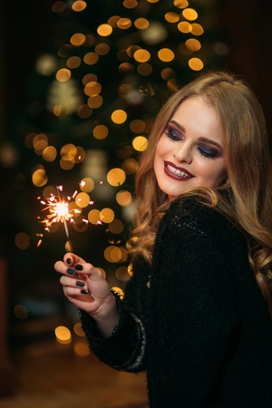 Portrait of young attractive celebrating woman holding sparkles. Background of christmas treeの写真素材