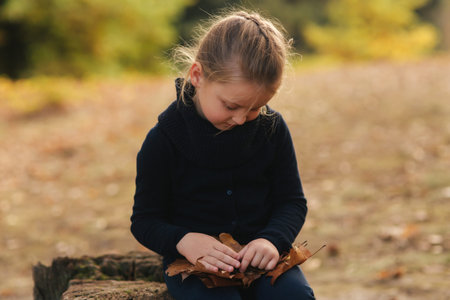 A little girl in black clothes is sitting on a stump and playing with autumn leaves. Blond hair girl