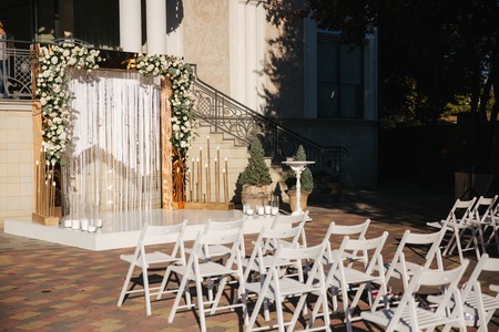 Weddind decoration on open air. Floral decor of a beautiful white and gold arch. Beautiful beckground view of restaurant and trees. Stool for ceremonyの写真素材