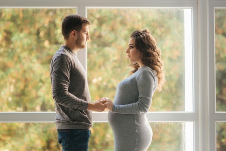 Happy family man and pregnant woman stand in front of big window in their house. They wait for a babyの写真素材