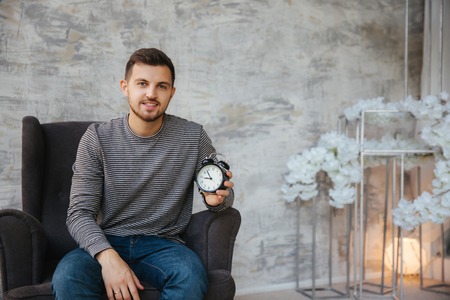 A young man sits on a chair and hold a clock in his hands. Time to stant. Handsome man smile and waiting suitable timeの写真素材