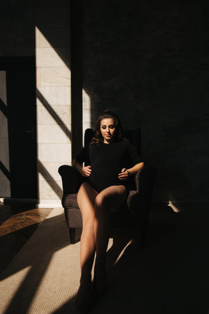 Beautiful pregnant woman sit on the chair. Sun rays fall on woman. She is dressed in black body suit