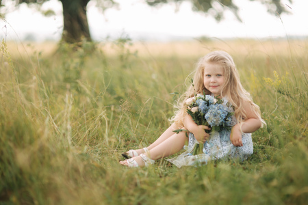 Little girl in sky blue dress with bouquet stand in field in front of big tree. Child smile and have funの写真素材