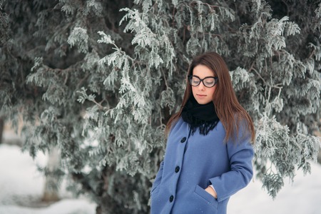Beautiful young woman with black glasses wearing winter coat color Blue Cobalt and black scarf covered with snow flakes. Background of white treeの写真素材