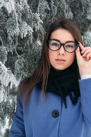 Portrait of happy young woman wearing scarf and blue coat stands next to the Christmas tree. black glassesの写真素材