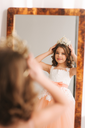 Little beautiful girl with brown hair in a Peach-colored dress. Girl try on diadem. Happy little princess look in to the mirror.の写真素材